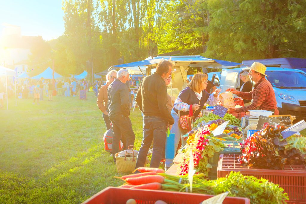 Farmers market : clients discutent avec un vendeur sous tente, produits frais et légumes exposés en plein soleil.