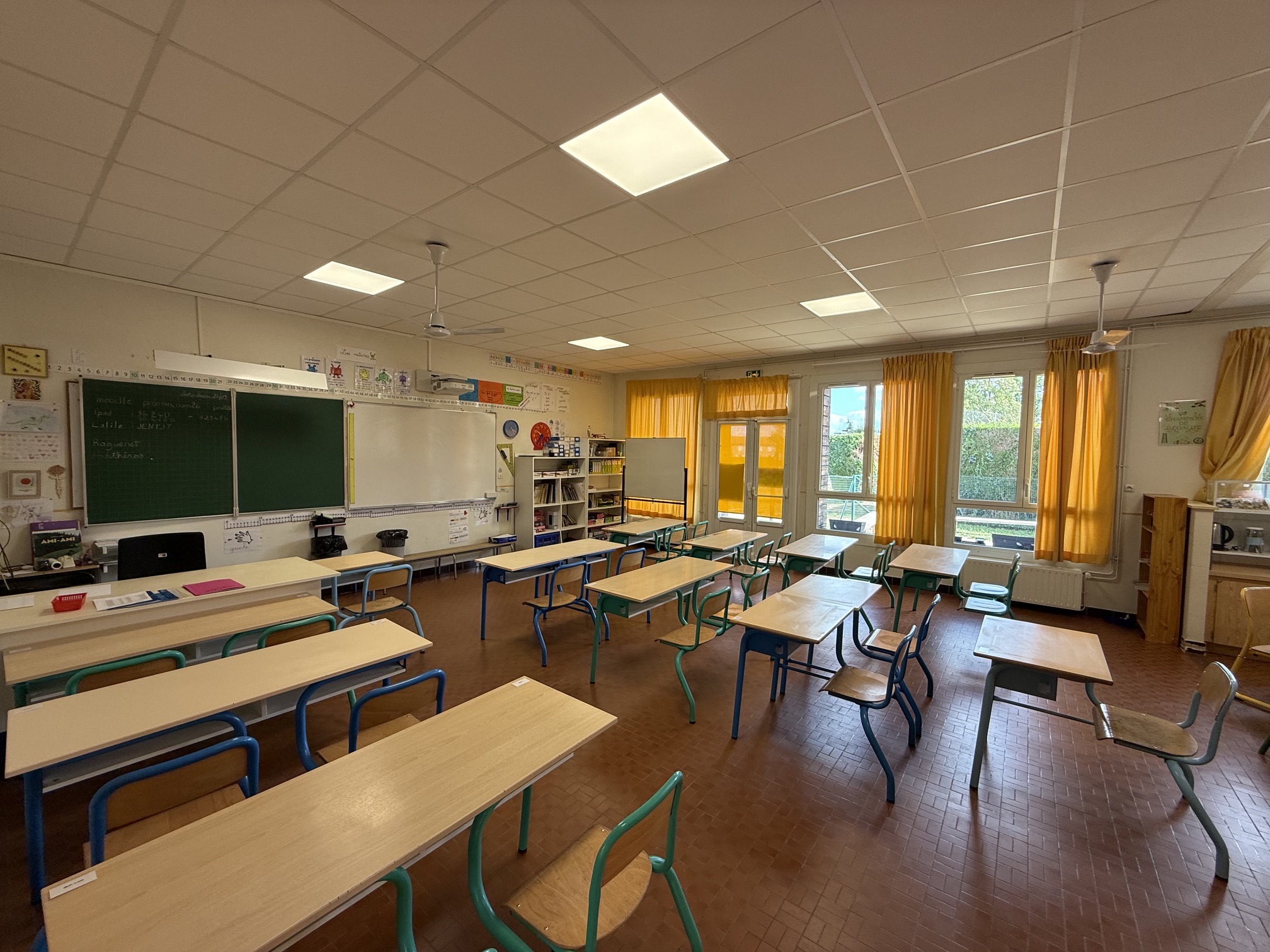 Salle de classe vide avec tables, chaises, tableau noir et fenêtres, éclairée par des néons.
