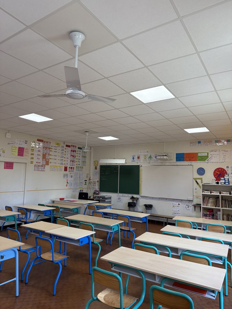 Salle de classe vide avec tables, chaises, tableau noir et affiches pédagogiques sur les murs, plafond avec éclairage et ve