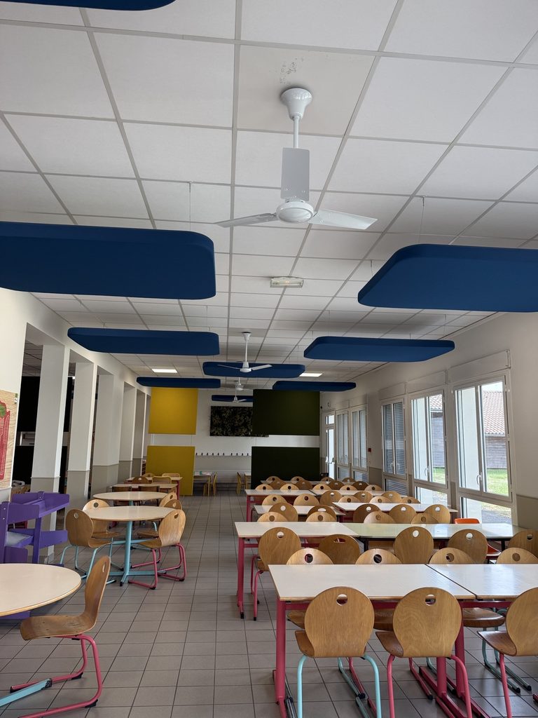 Salle de classe ou cantine avec tables, chaises et ventilateurs au plafond, éclairage moderne et murs colorés.