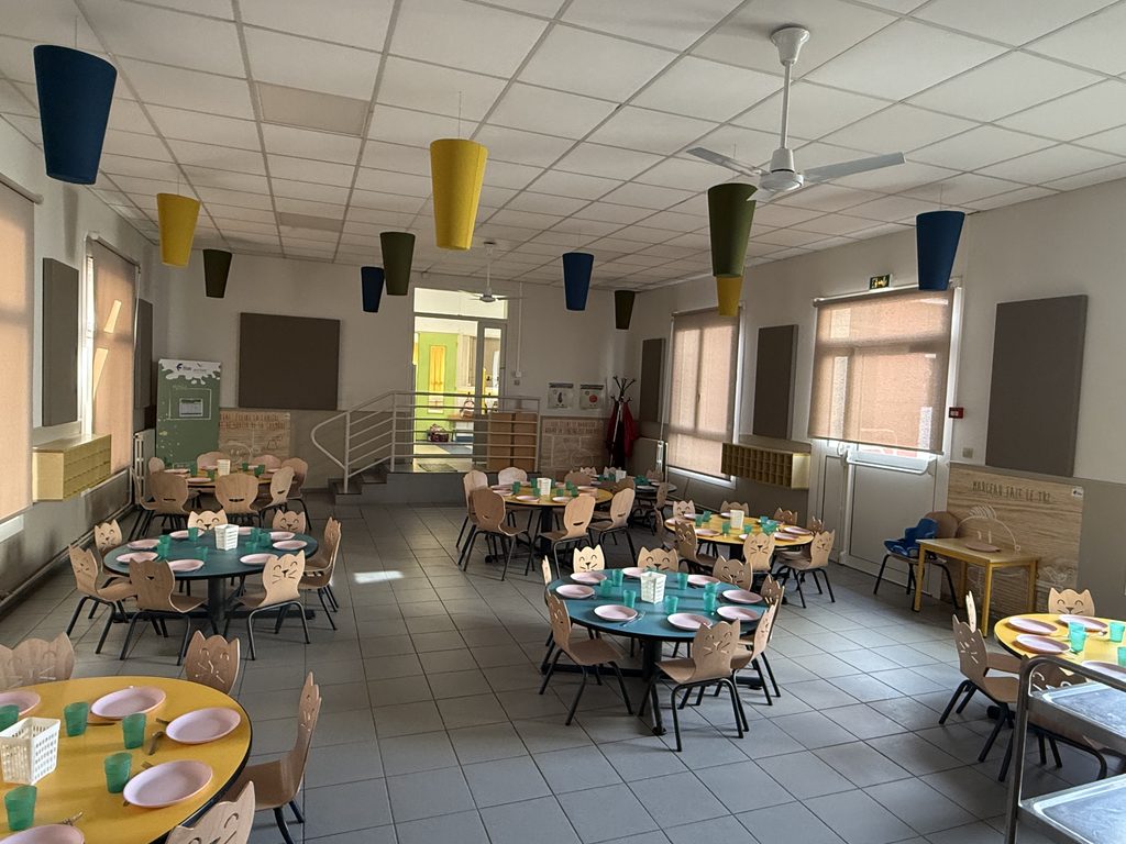 Salle de cantine enfantie avec tables rondes, chaises à forme d'animaux, couverts posés, luminaires colorés et espace aér