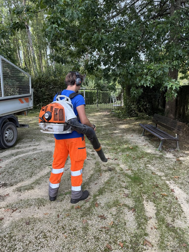 Un employé en tenue orange utilise une souffleuse dans un parc, équipé de protections auditives et d’un sac à dos.