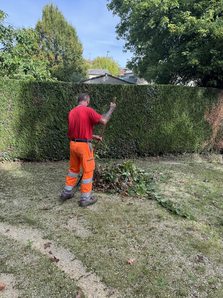 Un jardinier en tenue orange et rouge taille une haie avec un coupe-bordure dans un jardin bien entretenu.