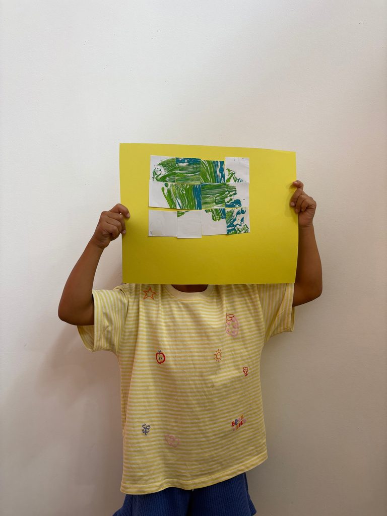 Enfant masqué par une feuille jaune tenant un dessin vert, chemise à rayures avec motifs colorés.