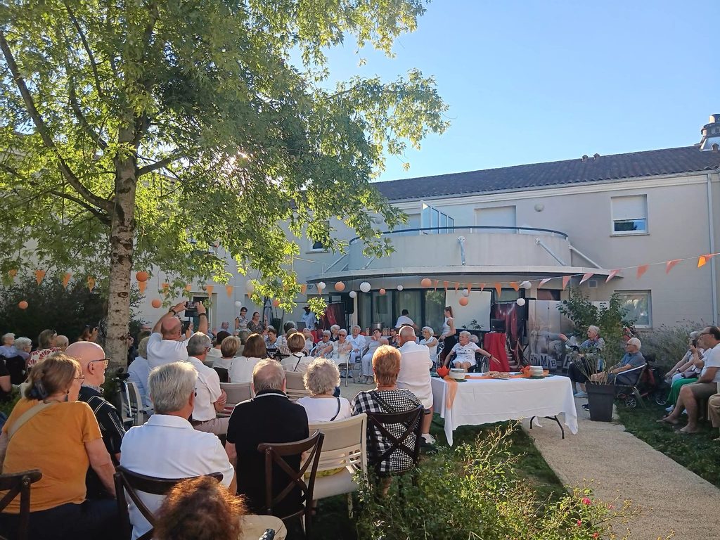 Groupe d’aînés assis en extérieur lors d’un événement festif sous des ballons colorés, en plein jour.