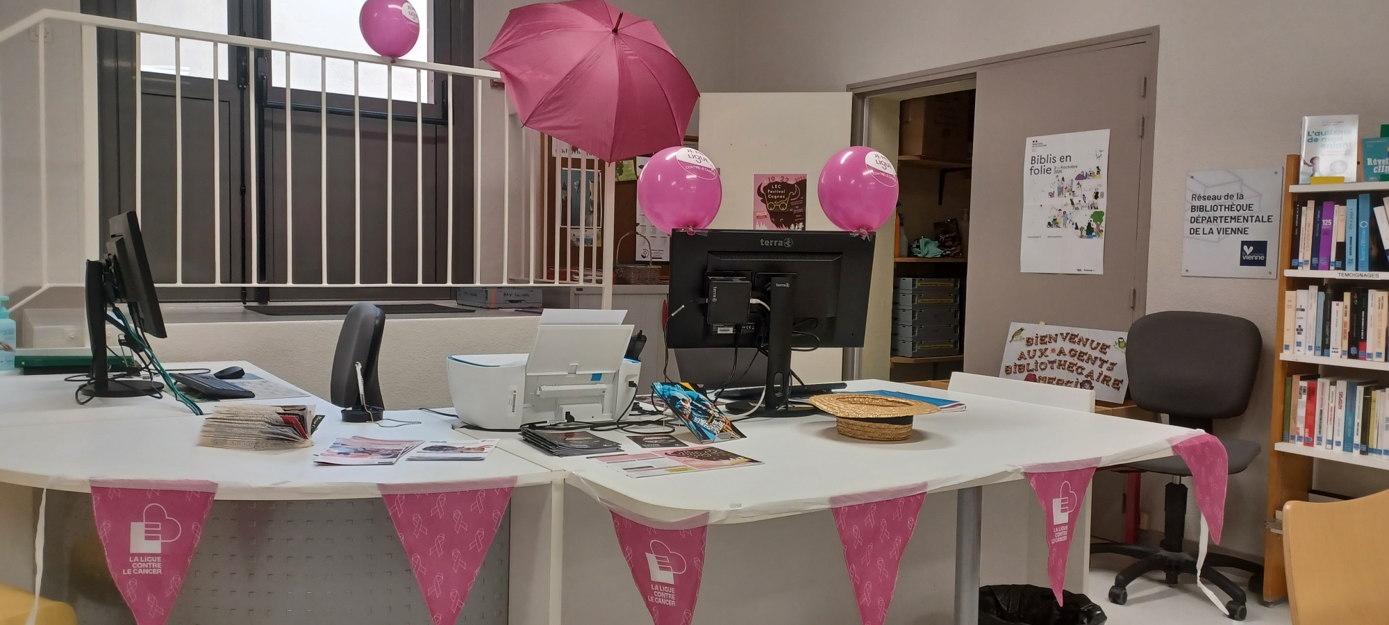 Poste de travail festif avec ballons roses, table décorée, écrans et bannières "La Ligue Contre le Cancer". Espace biblio