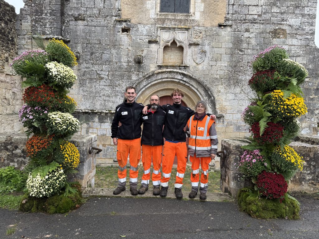 Quatre personnes en tenue orange devant une vieille pierre ornée de fleurs, devant une arche gothique.
