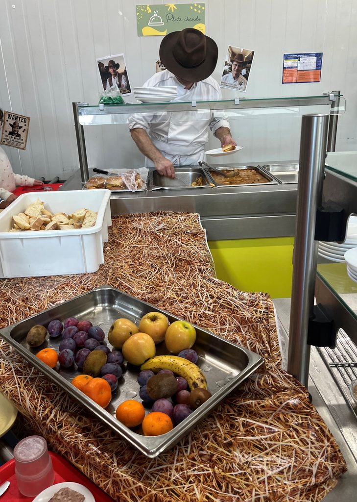 Un vendeur prépare des plats chauds derrière un comptoir avec fruits frais et pâtisseries en vitrine, ambiance rustique.