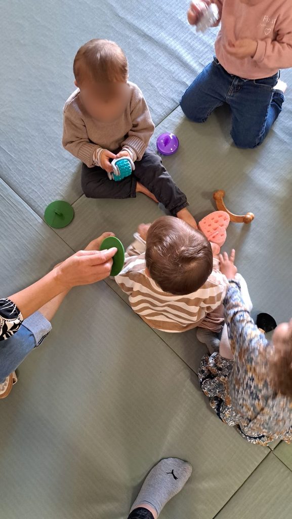 Deux bébés assis sur un sol avec jouets colorés, encadrés par des adultes, jouant ensemble.
