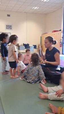 Enfants assis et debout écoutant une éducatrice en salle, activité ludique et éducative en groupe.