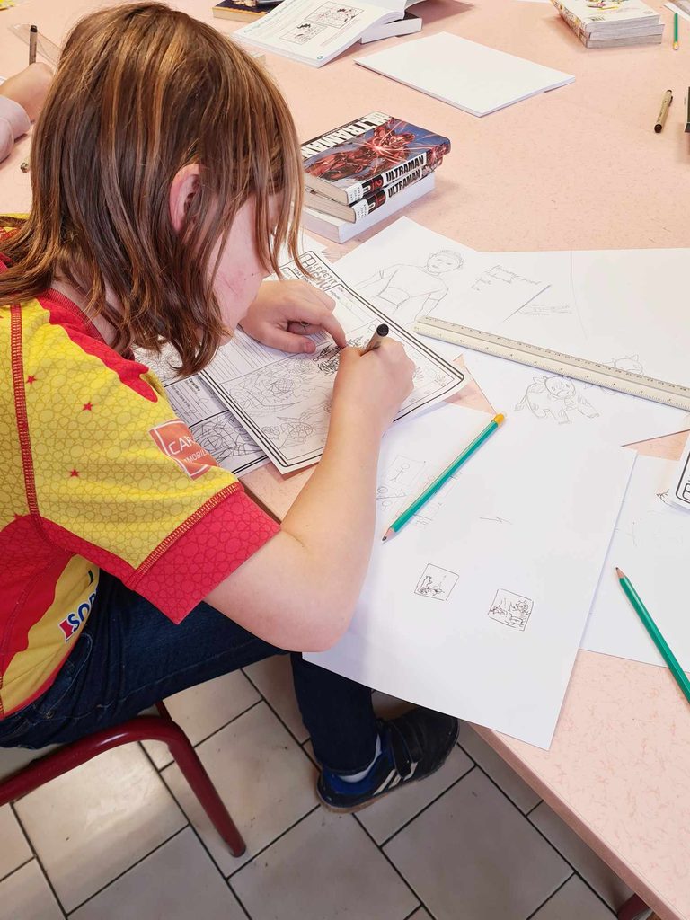 Enfant dessinant avec un crayon sur une feuille, entourée de livres et de matériel de dessin sur une table.