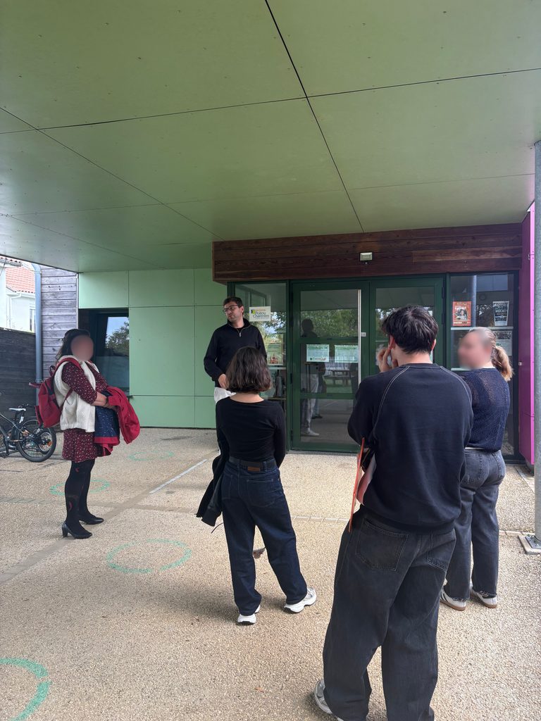 Un groupe de personnes attend devant une entrée verte, sous un auvent. Ambiance scolaire ou administrative.