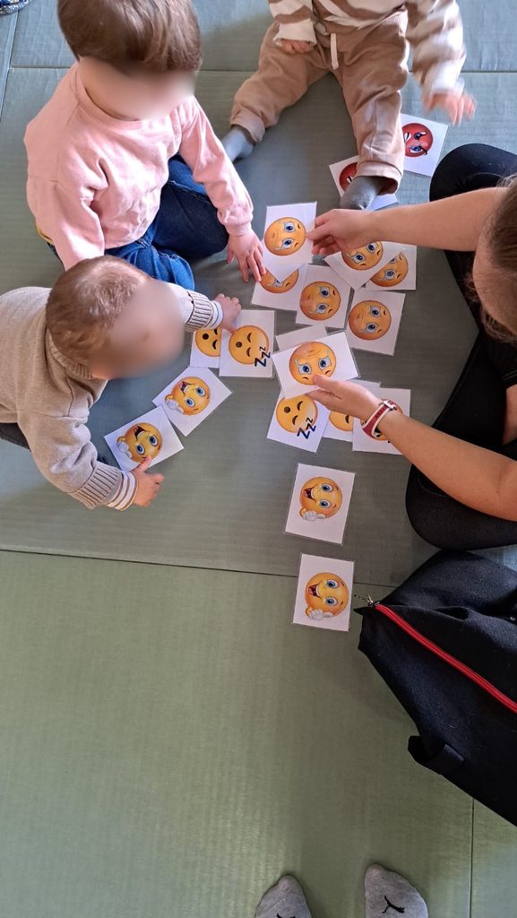 Trois enfants jouant avec une adulte à un jeu de cartes illustrées d’émojis sur une table basse.