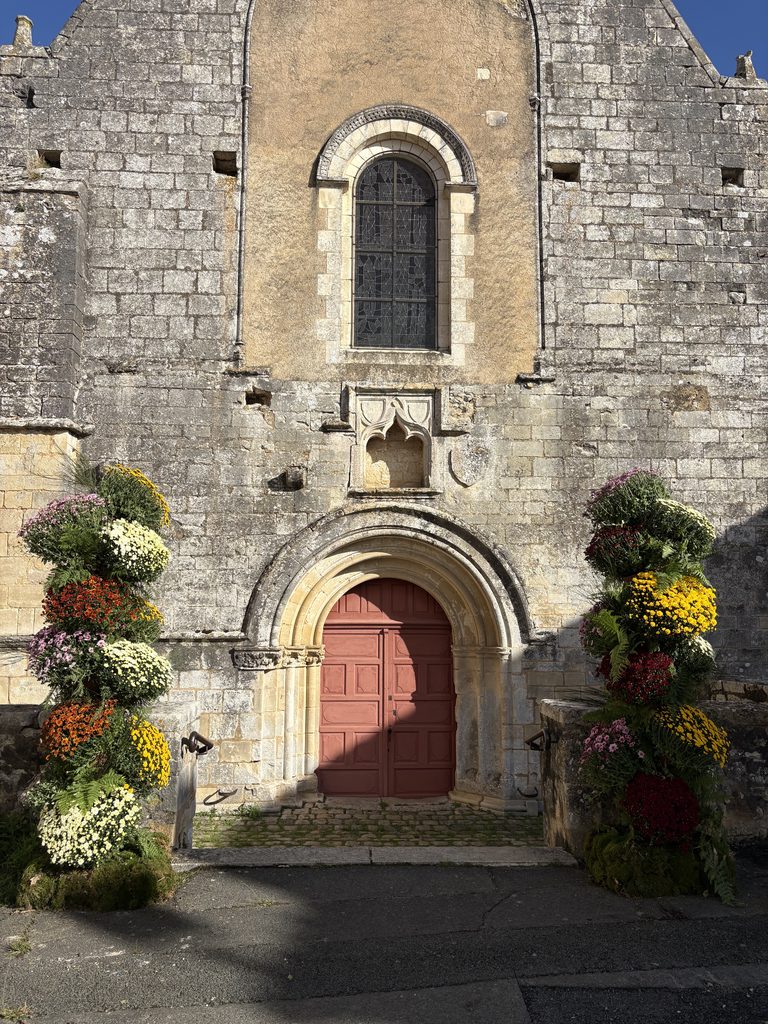 Ancienne église en pierre avec porte rouge, fenêtre gothique et fleurs colorées autour. Architecture historique et charman