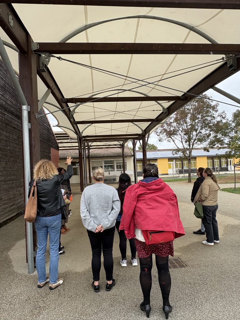 Groupe de personnes sous abri observant un bâtiment, vue de dos, extérieur scolaire couvert.