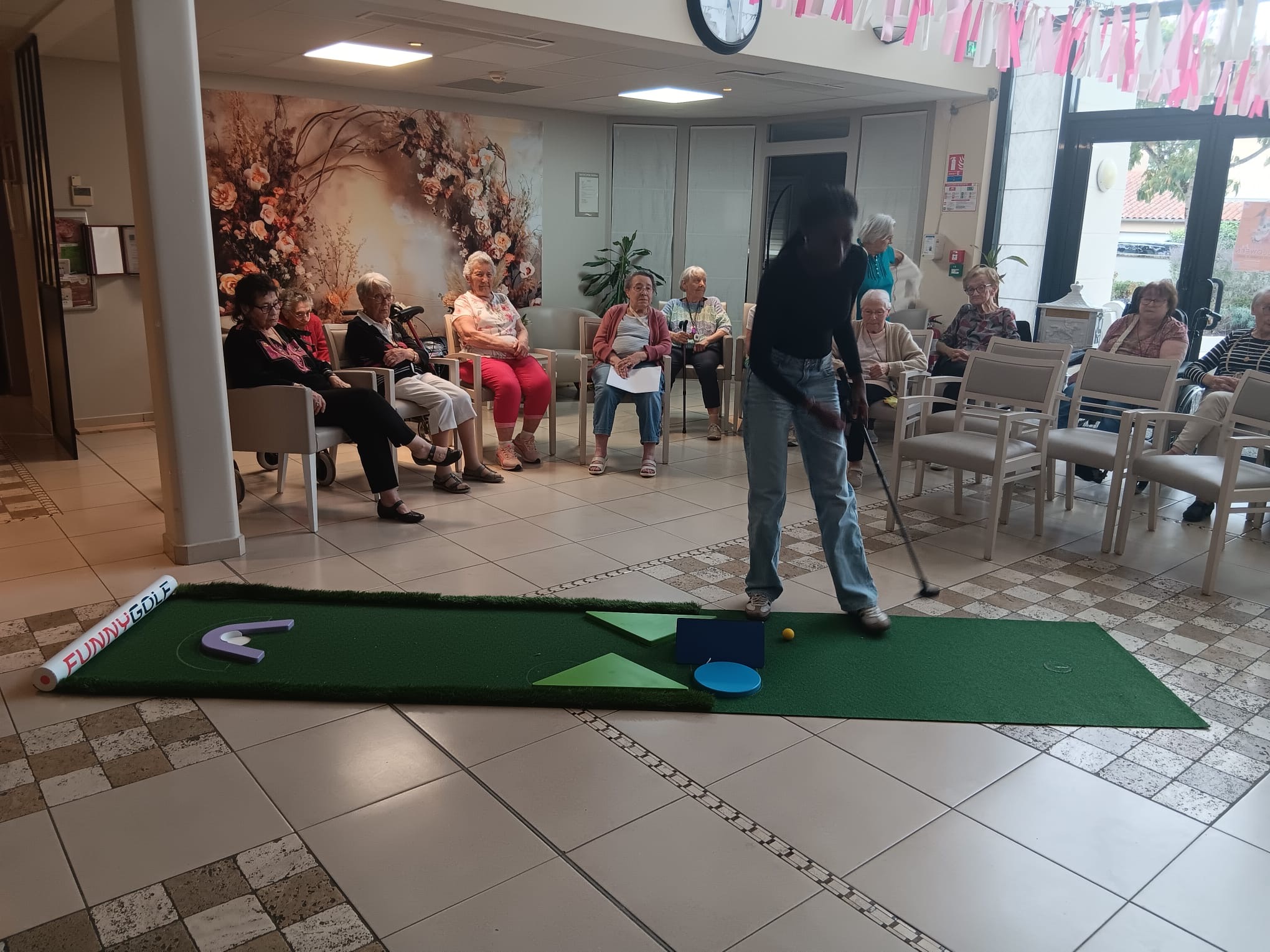 Un groupe de seniors assis observe une personne jouant au putting sur un tapis de mini-golf intérieur.