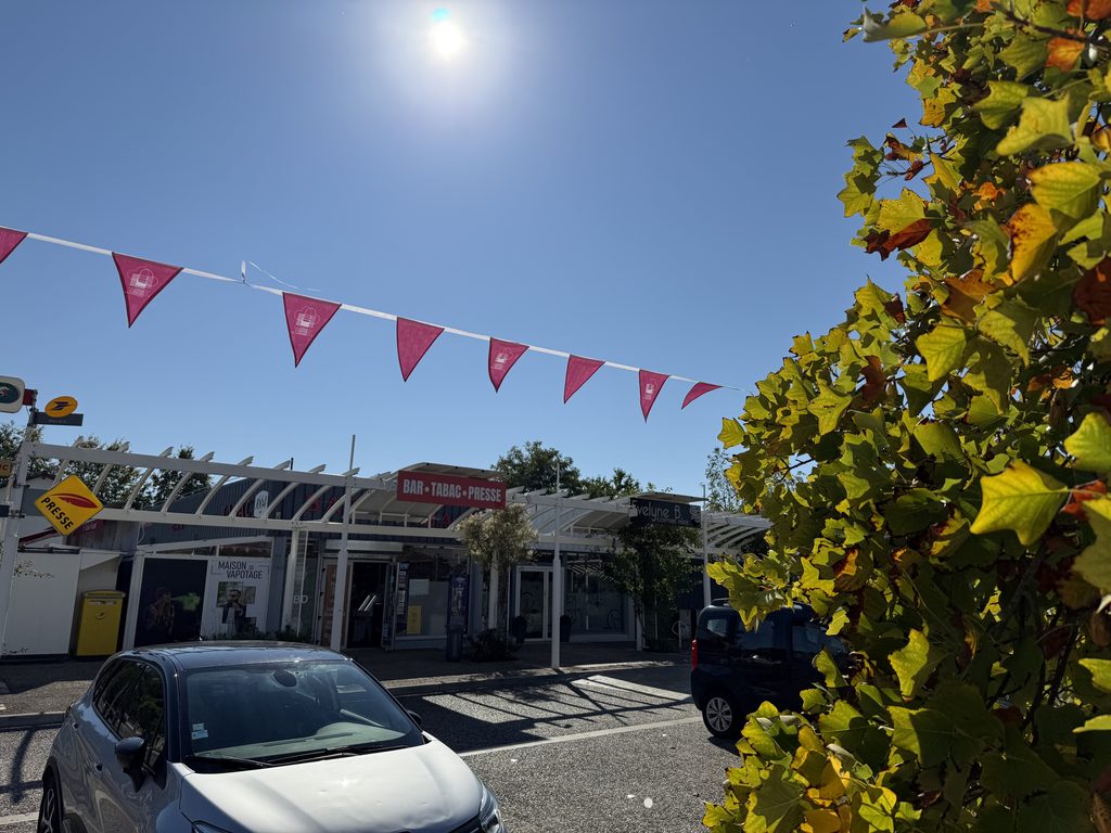 Pavillon avec drapeaux rouges, voitures et panneaux "bar-tabac-presse", sous un ciel bleu ensoleillé.