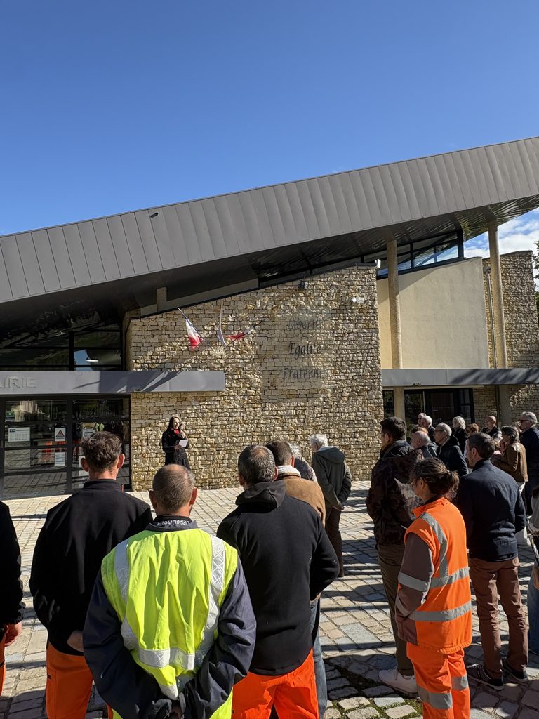 Groupe écoutant une personne devant un bâtiment en pierre, vêtus de vestes de sécurité orange et jaunes, extérieur enso