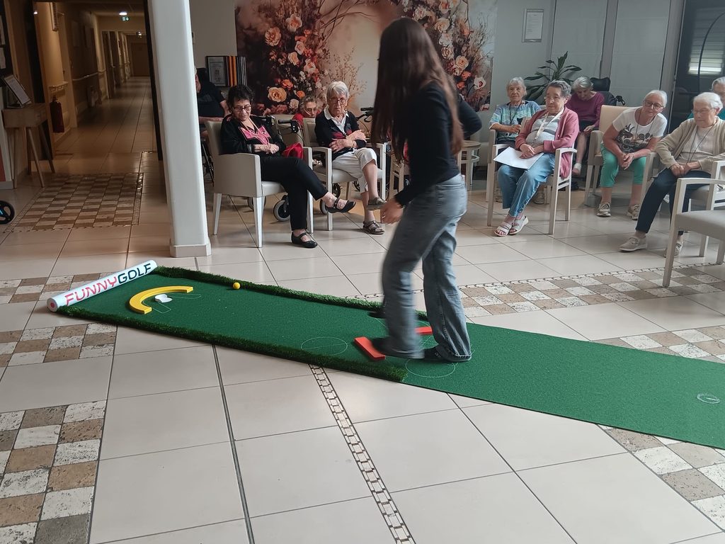 Personne jouant au mini-golf sur un tapis vert en intérieur, devant un groupe dʼobservateurs assis.