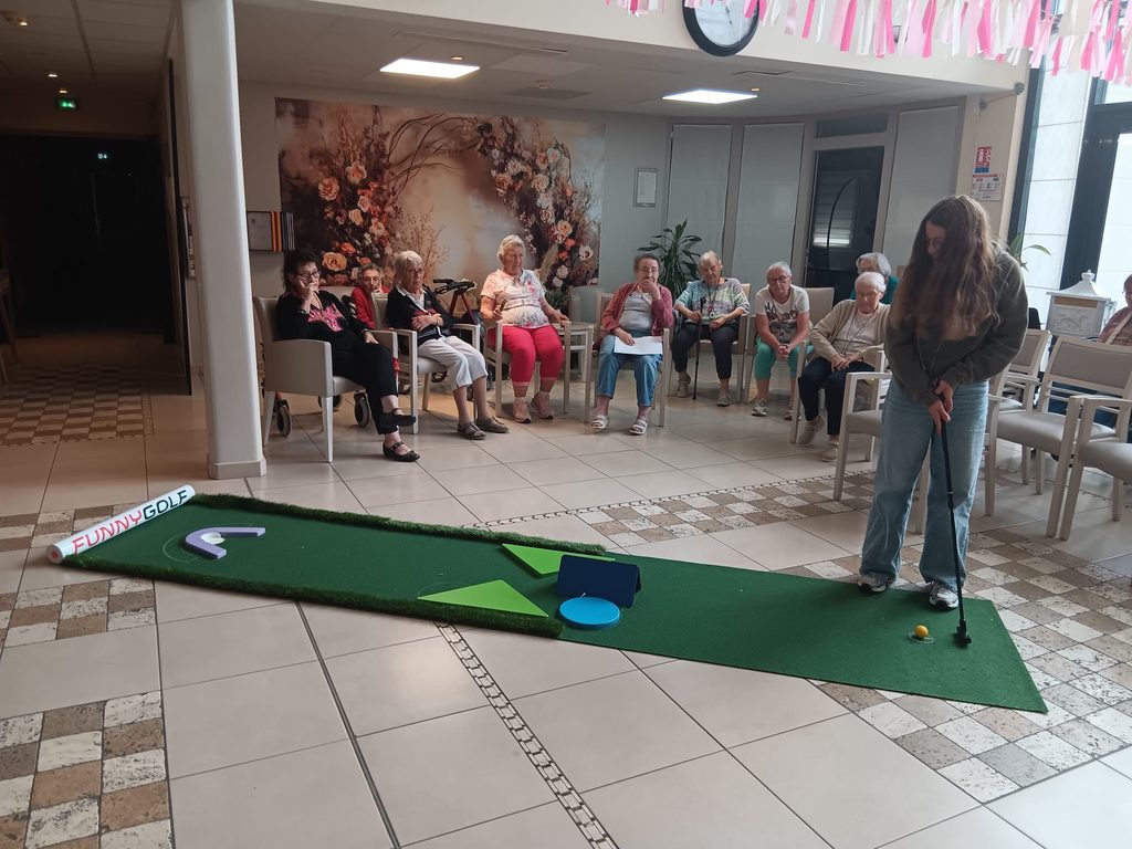 Un groupe d’aînés observe une personne jouant au mini-golf en intérieur, sur un parcours avec obstacles colorés.
