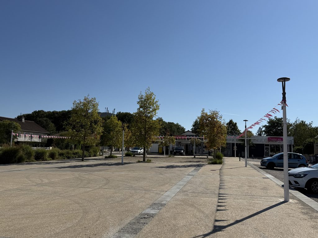 Parking ensoleillé avec trottoir, arbres et drapeaux rouges et blancs, voitures garées, bâtiments en arrière-plan.