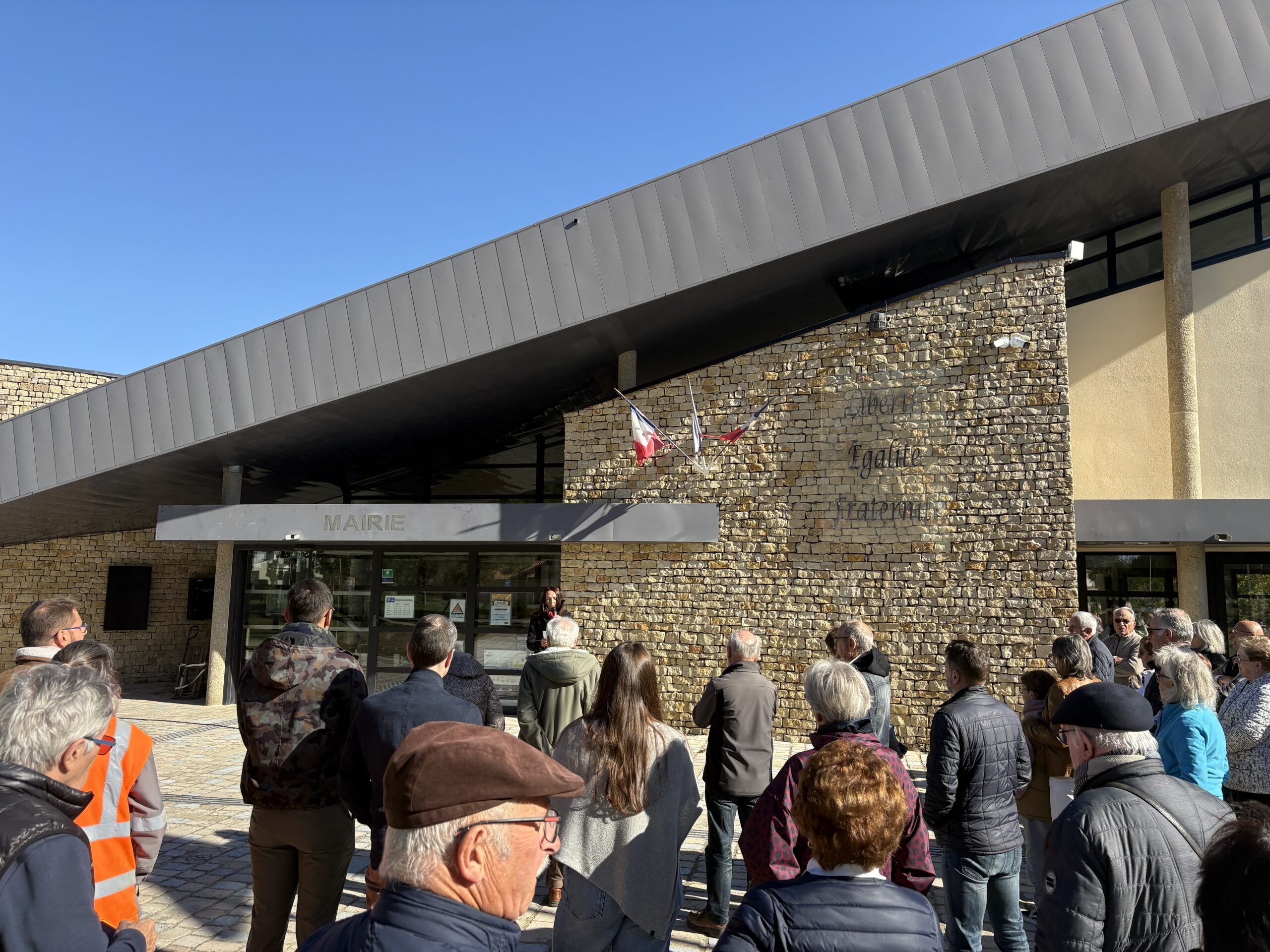 Groupe assemblé devant une mairie, discours en extérieur sous un auvent, bâtiment en pierre avec drapeaux français.