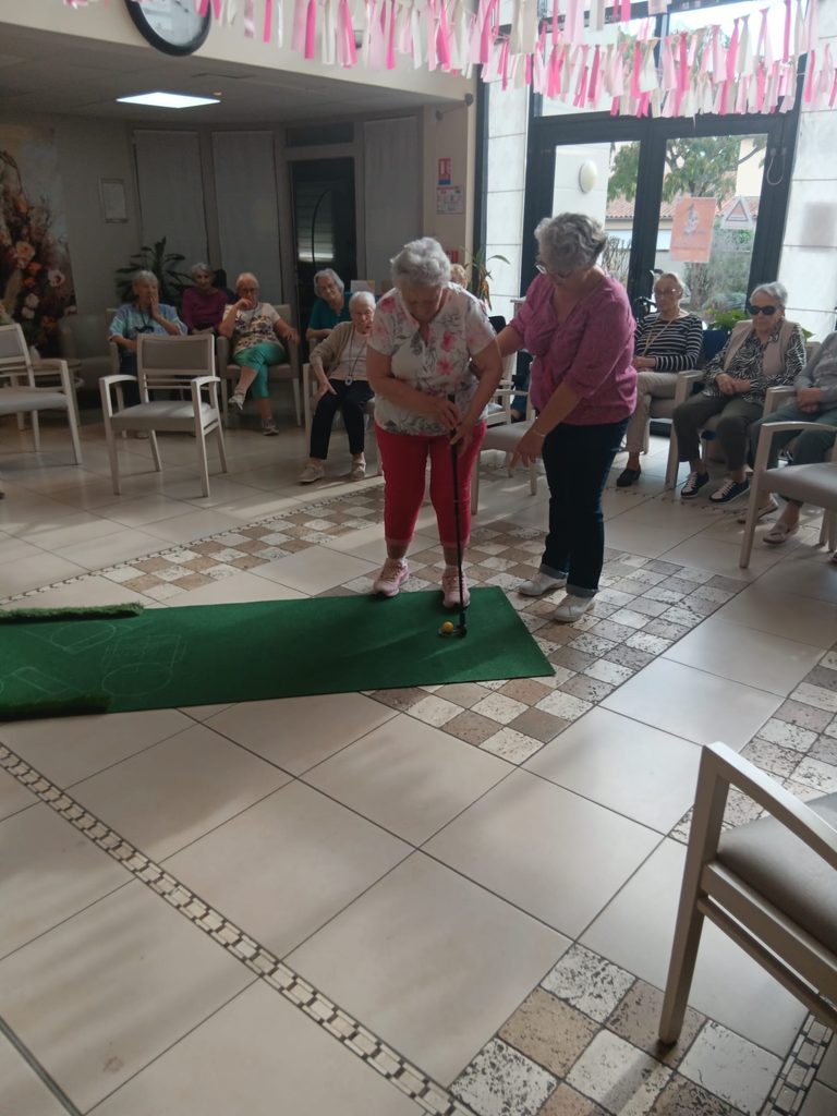 Deux femmes font de l'exercice sur un tapis vert, entourées de seniors assis observant dans une salle lumineuse.