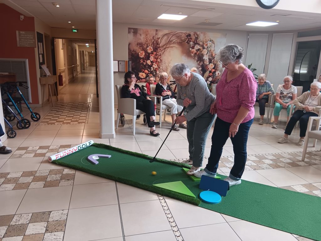 Deux personnes jouent au putting sur un tapis de golf miniature, entourées de spectateurs assises dans un hall d’accueil.