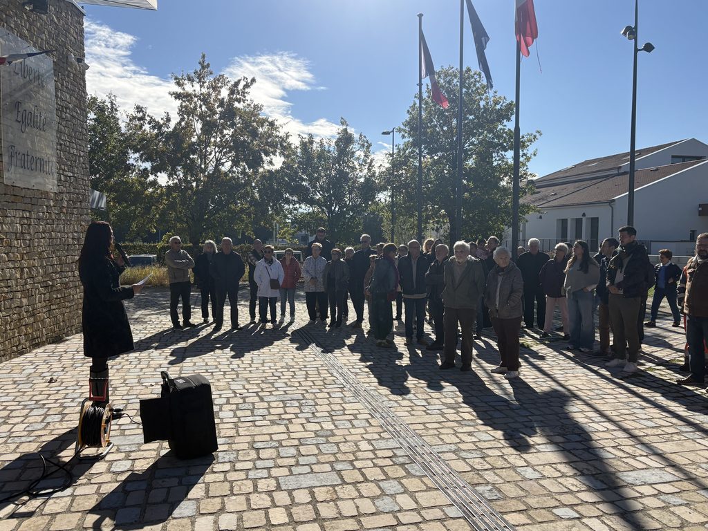 Groupe d’adultes en assemblée devant un monument, sous des drapeaux, en plein jour.