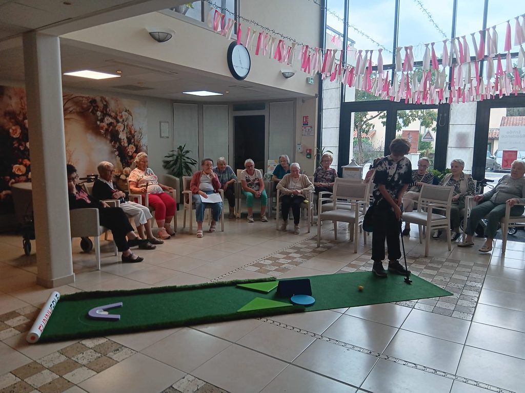 Groupe de seniors assis observant une personne jouant au mini-golf en intérieur, décoré de guirlandes colorées.