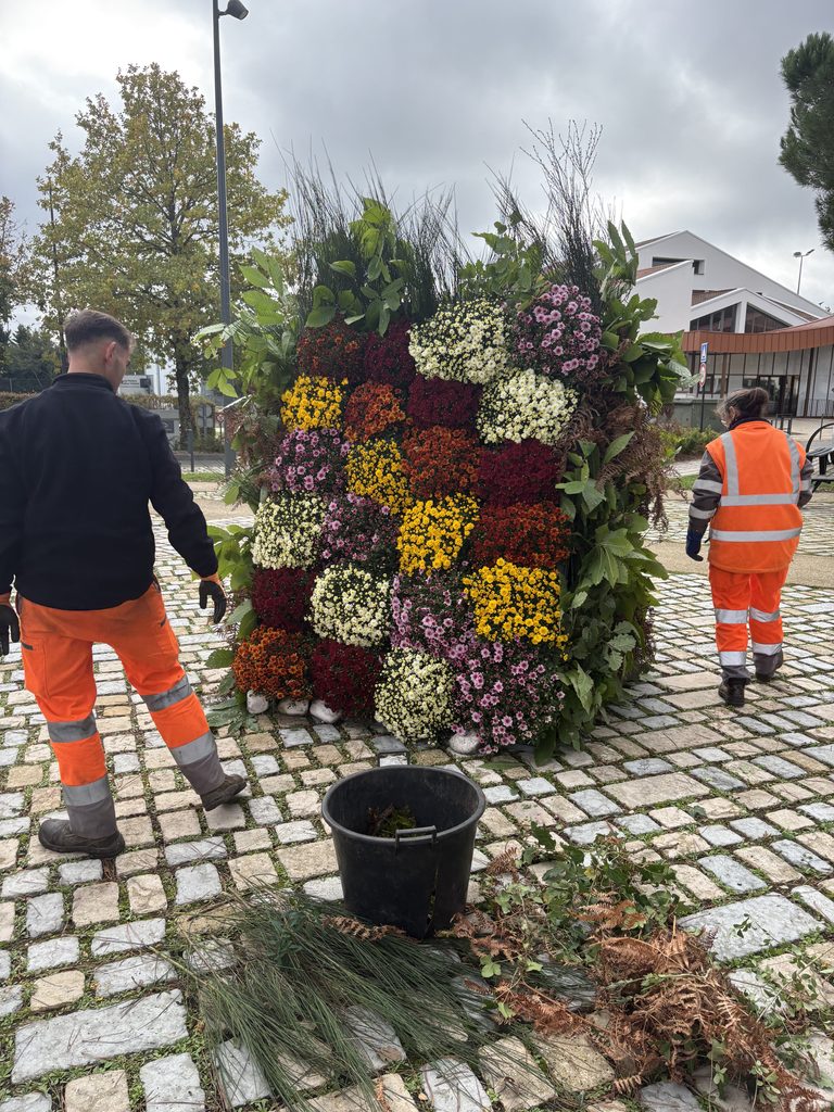 Deux travailleurs en combinaison orange ajustent une grande composition florale sur une place pavée.