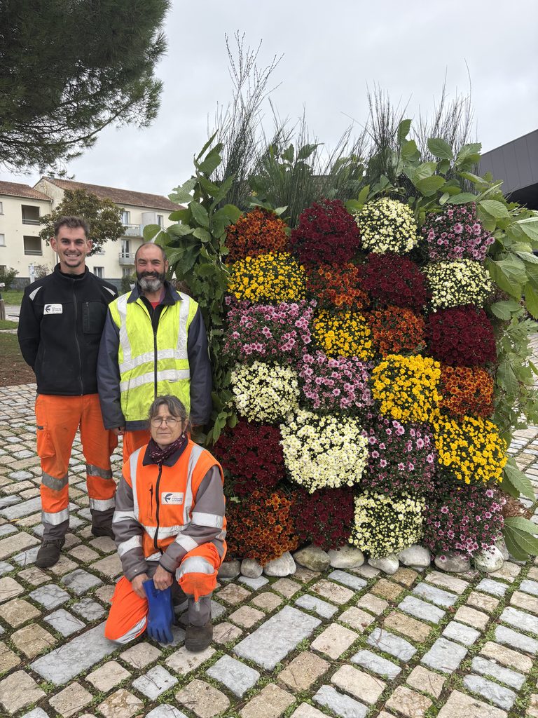 Trois jardiniers en tenue orange et jaune posent devant une haie fleurie colorée sur un sol pavé.