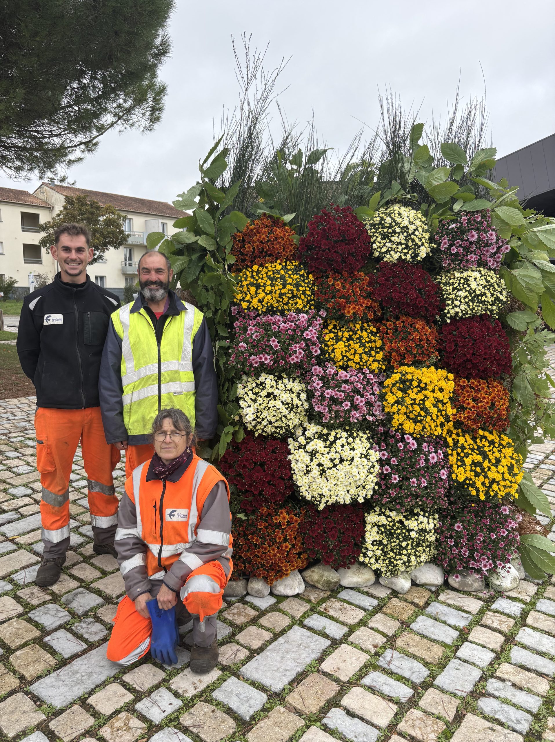 Trois jardiniers en tenue orange et jaune posent devant une haie fleurie colorée sur un sol pavé.